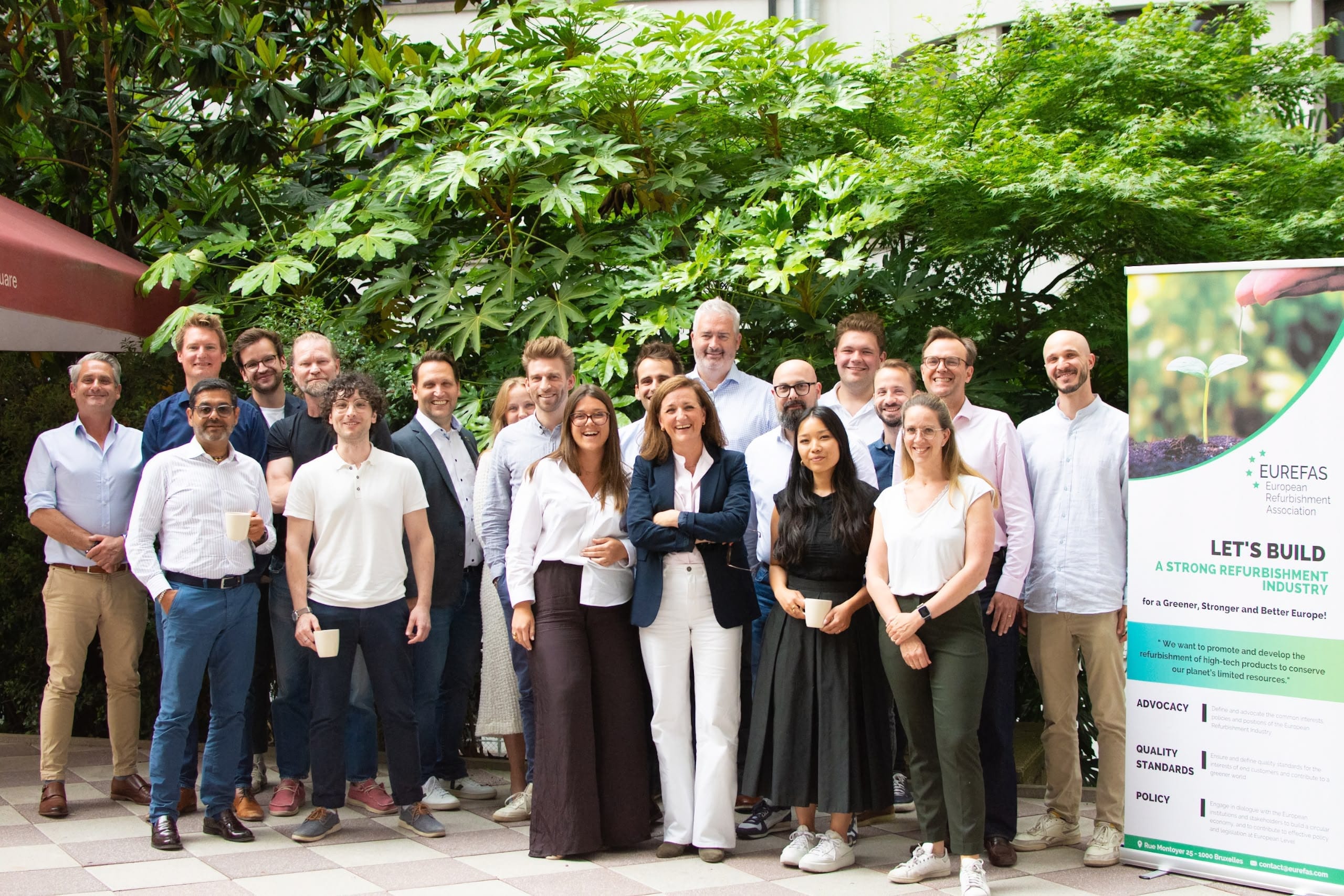Group photo of about 20 people standing outdoors on a tiled patio, smiling in front of dense green foliage; a roll‑up banner at right shows a sprouting plant image and text.