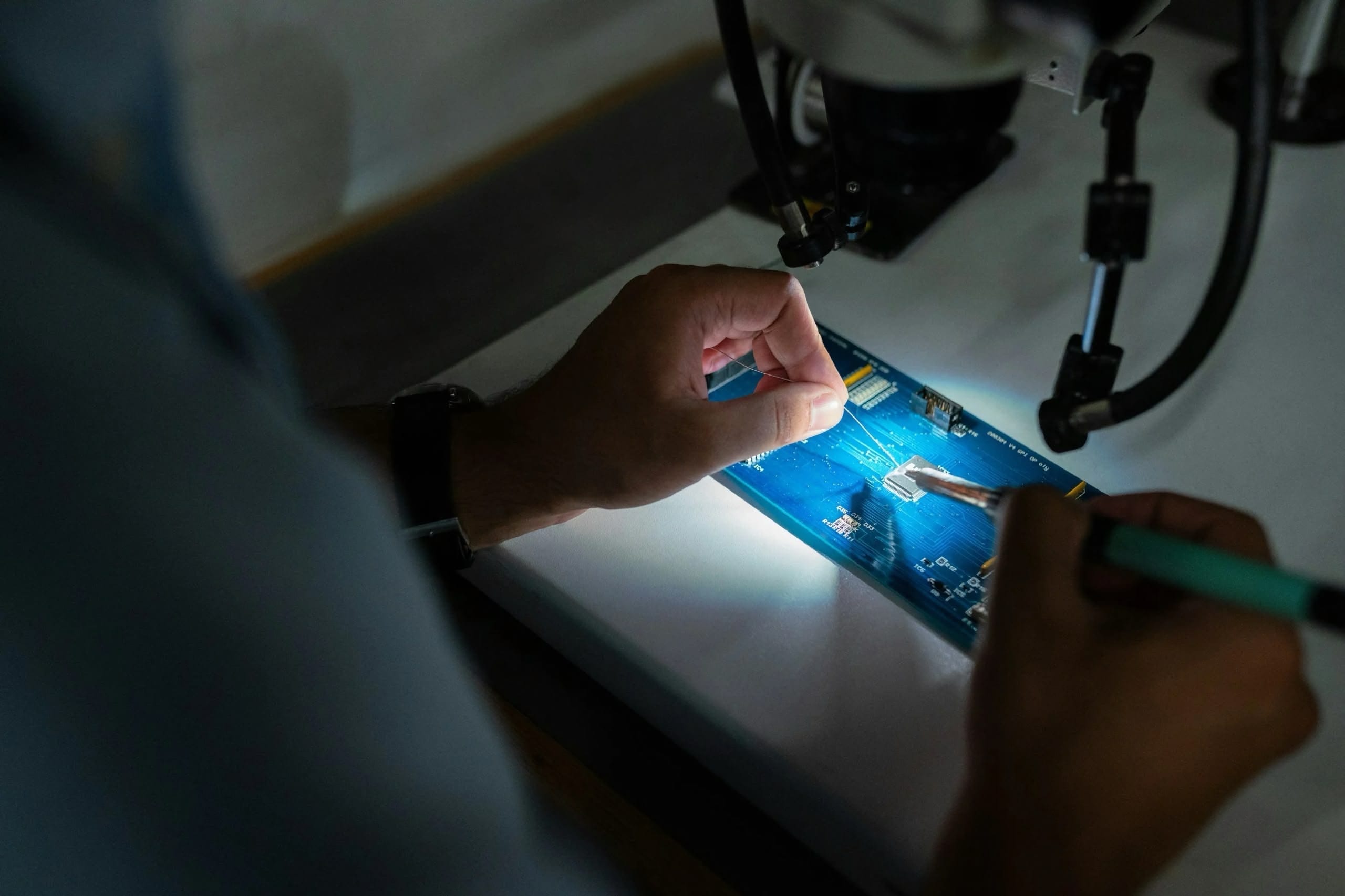 Close-up of hands working under a microscope on a blue printed circuit board; one hand positions fine wires while the other uses a precision tool, with a focused light illuminating the components.