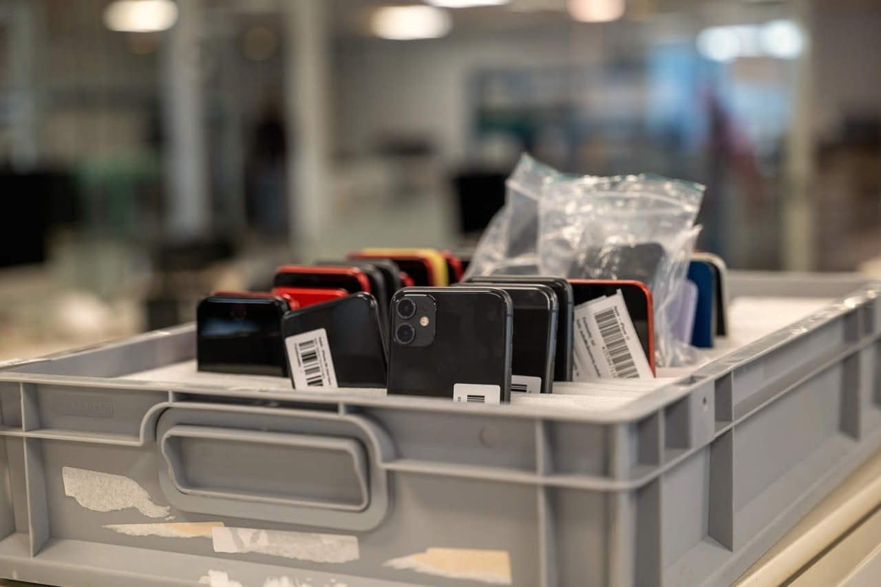 Gray tote filled with assorted smartphones standing upright with barcode stickers and a bag of accessories, in a warehouse or lab setting.